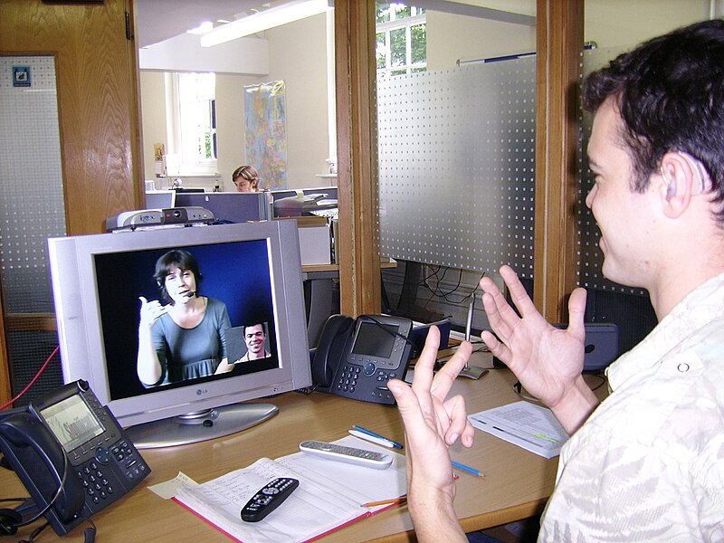 File:Deaf or HoH person at his workplace using a Video Relay Service to communicate with a hearing person via a Video Interpreter and sign language SVCC 2007 Brigitte SLI + Mark.jpg