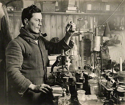 Photo of a man with a pipe in his mouth standing at a table with a microscope and numerous specimen vials