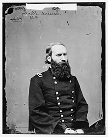 Serious man with short hair and a full, long beard in army uniform, sitting and looking to the right.