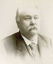 Ein kahlköpfiger Mann mit Schnurrbart Ende sechzig.  Er trägt einen schwarzen Mantel, ein weißes Hemd und eine gestreifte Krawatte