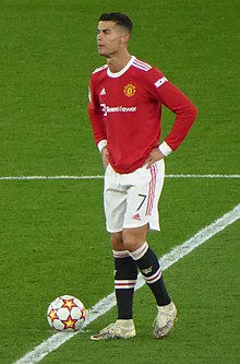 A imagem mostra Cristiano Ronaldo, homem, branco, com 1,88m, com uma camiseta vermelha e um calção branco, no centro do campo, com as mãos na cintura.