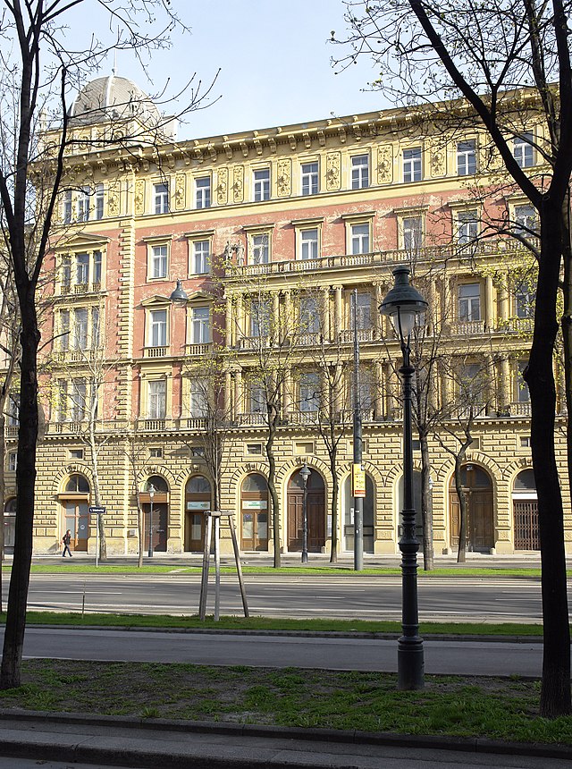 Frontale Farbfotografie einer Gebäudefassade mit fünf Stockwerken. Im Erdgeschoss bestehen die Türen und Fenster aus Rundbögen. Der Mittelteil des ersten und zweiten Stockwerks ist mit Säulen verziert und auf einem Balkonende stehen zwei Figuren. Zwischen den Fenstern im Obergeschoss befinden sich Ornamente und auf der linken Dachseite ist eine Kuppel. Der Vordergrund ist von einer Straße und kahlen Bäumen durchzogen.