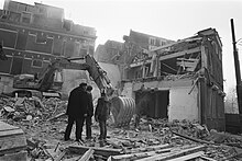Three men stand in front of arm of mechanical digger being used to demolish buildings