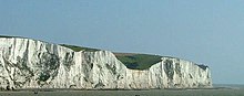 Uma fotografia panorâmica de uma seção das falésias brancas de Dover, com o canal da Mancha em frente