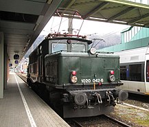 Bild der in grüner Farbe lackierten Museumslok 1020.42‑6, die im Bregenzer Hauptbahnhof auf die Weiterfahrt wartet. Im Hintergrund ist der Nahverkehrstriebwagen 4024 der ÖBB zu sehen.