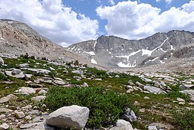 A granite ridgeline mottled with snow with a pronounced dip on the left marks Mather Pass, rising above a green alpine landscape.