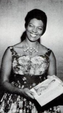 A smiling African-American woman, wearing a sleeveless floral dress and a necklace, holding a book or a box in both hands