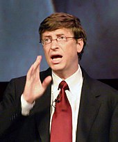 Head and shoulders portrait of talking 50-year-old man in rimless glasses, business suit, and power red tie, with raised right hand gesturing.