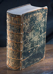 Photographie en studio d'une très vieille Bible debout verticalement sur une surface en bois avec la colonne vertébrale tournée aux trois quarts vers le spectateur.  La couverture est en cuir noir et est craquelée et usée.