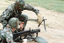 The M60 machine gun crew member responsible for hot barrel changes uses protective asbestos gloves to prevent burns to the hands.