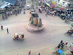 Автобусная остановка Madhupur, тройное соединение трех важных дорог Бангладеш. Три дороги ведут в Тангайл, Мименсингх и Джамалпур соответственно.  Ананасы обозначают область выращивания ананасов вокруг Мадхупура.