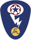 Oval shaped shoulder patch with a deep blue background. At the top is a red circle and blue star, the patch of the Army Service Forces. It is surrounded by a white oval, representing a mushroom cloud. Below it is a white lightning bolt cracking a yellow circle, representing an atom.