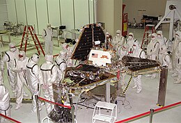 A group of scientists, all wearing white protective clothing, gather around a spacecraft as it's being folded into its launch position; a triangular pyramid shape.