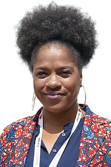 Fotoporträt einer schwarzen Frau, die direkt in die Kamera blickt. Sie trägt ein bunt gemustertes Oberteil, die Haare sind in einer Afrofisur hochgesteckt.