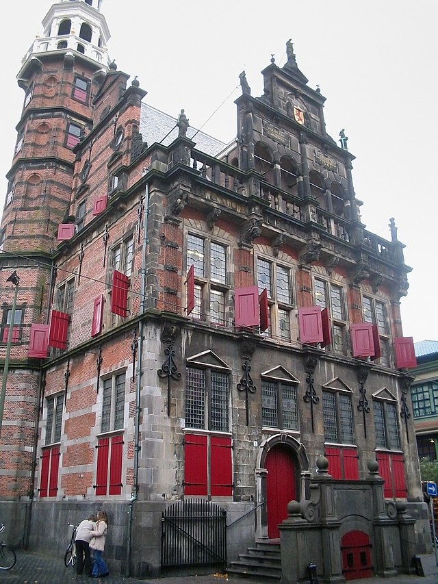 Seitliche Farbfotografie eines zweigeschossigen, braunen Gebäudes mit zwei Dachfassaden an beiden Seiten, die mit Figuren und Reliefs ausgestattet sind. An einigen Fenstern hängen rote Fensterläden und der rote Rundbogeneingang ist durch Stufen erreichbar. Im Hintergrund befindet sich ein Turm mit einer weißen Spitze.