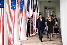 Donald Trump e Amy Coney Barrett camminano fianco a fianco lungo il West Wing Colonnade; Le bandiere americane pendono tra le colonne alla loro destra