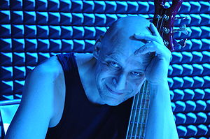 bald White male wearing a dark tank top, holding a six-string bass guitar in a blue-lit room, smiling at camera