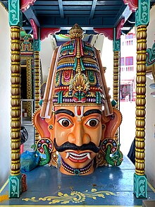 A big moustached male head, with big eyes, big ears and thick eyebrows. Fangs protrude from the sides of his mouth. The head wears a conical crown, with a cobra hood at the top. A floral garland and gold necklace are seen around the neck.