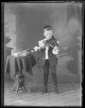 A “young boy playing the violin.” Beside him is a table with likely a banjo on it.