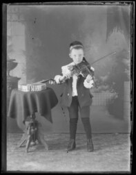 A “young boy playing the violin.” Beside him is a table with likely a banjo on it.