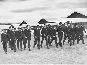 Fifteen men in dark military uniforms walking in front of biplanes and huts