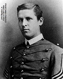 A black and white picture of John Bigelow Jr., a young white male in his United States Military Academy Cadet uniform. His Cadet uniform has a starched white collar and his tunic has large round brass buttons interconnected by horizontal braiding. He is clean shaven with neatly combed short dark hair parted to his right. His right sleeve shows two stripes indicating he is a Cadet Lieutenant.