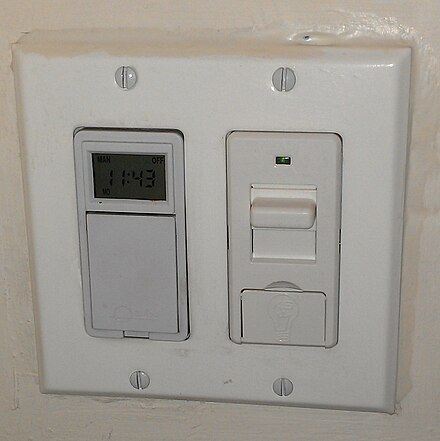 A KosherSwitch unit shown installed alongside a traditional Shabbos clock/timer.  The switch is in Sabbath Mode, and may be used, since a green status light is visible.