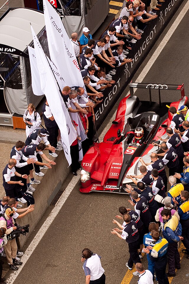 A red, black and white sports prototype being driven down a pit lane with people applauding the driver