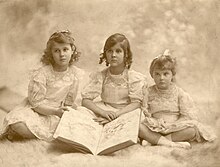 Trois petites filles assises en tailleur avec, devant elles, un grand livre.