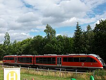 Zweiteiliger, roter Zug vor einem Wald. Unten rechts das Wappen