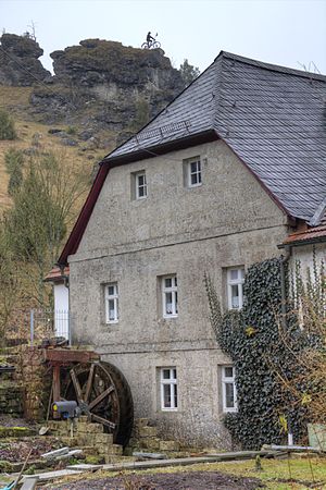 Blick auf den Nordgiebel der Stoffelsmühle. Davor das 2011 installierte Wasserrad; oberhalb der Mühle die Radfahrerstatue von Kleinziegenfeld