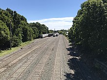 A fairly small railroad yard is seen from a bridge that crosses above it. It contains about 7 to 8 tracks, and parked groups of freight cars occupy several of the tracks.