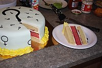 A white frosted cake decorated with black question marks and Mars and Venus symbols; the cake has been cut open and a piece sits on its side on a paper plate to the right. There are three layers to cake; the top and the bottom layers are off-white and the middle layer is pink.