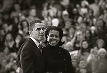 Barack en Michelle Obama, droeg Een Donkere outdoor Kleding, ten overstaan van Een menigte.  Zijn uitdrukking is gedempt;  Ze Heeft Een Brede Glimlach.