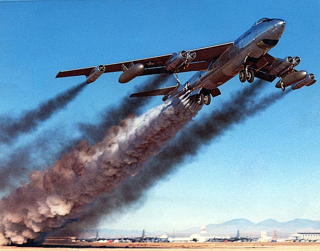 A shiny needle-nosed four-engine jet aircraft takes off. It is trailing an enormous amount of smoke.