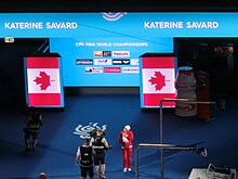 Farbfoto von Katerine Savard im roten Trainingsanzug mit weißer Bademütze. Sie ist nur klein im Vordergrund zu sehen, im Hintergrund eine riesige Medienwand mit den Namen diverser Sponsoren eingerahmt von der kanadischen Flagge.