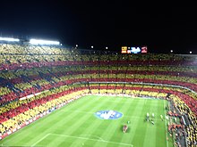 Camp Nou. The home fans of FC Barcelona are creating a mosaic of the Catalan flag before El Clasico. The top right corner of the club's crest also features a Catalan flag. Camp Nou during El Clasico October 2012.jpg