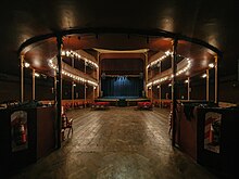 Vista de la sala del teatro Margarita Xirgu desde la entrada a la misma. PIso de madera y pocas butacas rojas. De fondo, en el escenario, un telón azul.