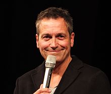 Portraitfoto von Dieter Nuhr, lächelnd mit Mikrofon vor dem Mund. Darunter seine Unterschrift