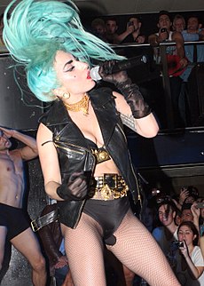 Gaga performing onstage wearing black leather jacket and bodysuit. She has blue hair