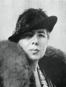 Black and white portrait photograph of Beatrice Gordon Holmes. Framed so we can only see her from the shoulders upwards, she is turning to look into the camera - a middle-aged woman wearing a black hat, with a large fur draped around her shoulders, and bearing a resolute expression on her face.