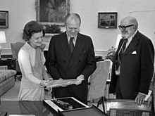 Černobílá fotografie, na které jsou tři postavy v místnosti. Prezident Gerald Ford a první dáma Betty Fordová si společně s Adamsem prohlížejí fotografie.