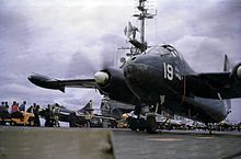 Large black twin-propeller aircraft on the deck of an aircraft carrier. The island is in the background and other aircraft are parked astern. Various crewmen are standing around, wearing brightly colored jerseys.