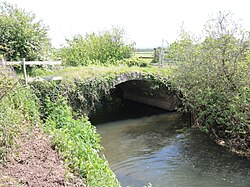 The bridge is shown spanning the river that is bordered by embankments covered in grass. There is shrubbery in the background and the parapet is covered in grass and weeds. Wooden fencing is visible to the left of the image.