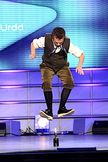 Photo d'un homme en habit traditionnel gallois, en plein saut au-dessus d'une bougie sur une scène, les jambes pliées et le regard tourné vers le bas. Un éclairage néon éclaire la scène en arrière-plan.