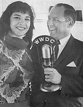 A young woman with long dark hair standing beside an older man in a jacket and tie. The man is holding up a large microphone.