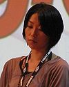A photograph of a woman with short black hair facing left and looking down while wearing a black lanyard around her neck that reads "I WANT ANIMAX"