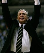 Koyu yeşil ve gümüş kravatlı siyah bir takım giyen Pat Riley, iyi bir oyunu kutlarken ellerini havaya fırlatıyor.