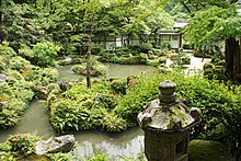 Photo couleur d'un jardin paysager japonais comprenant des massifs de verdure, des arbres, des rochers, un étang et, au premier plan, une lanterne de pierre (couleur domiante : vert).