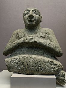 Statue af mørk sten, der repræsenterer bysten på en karakter med foldede hænder, og som ruller øjnene.